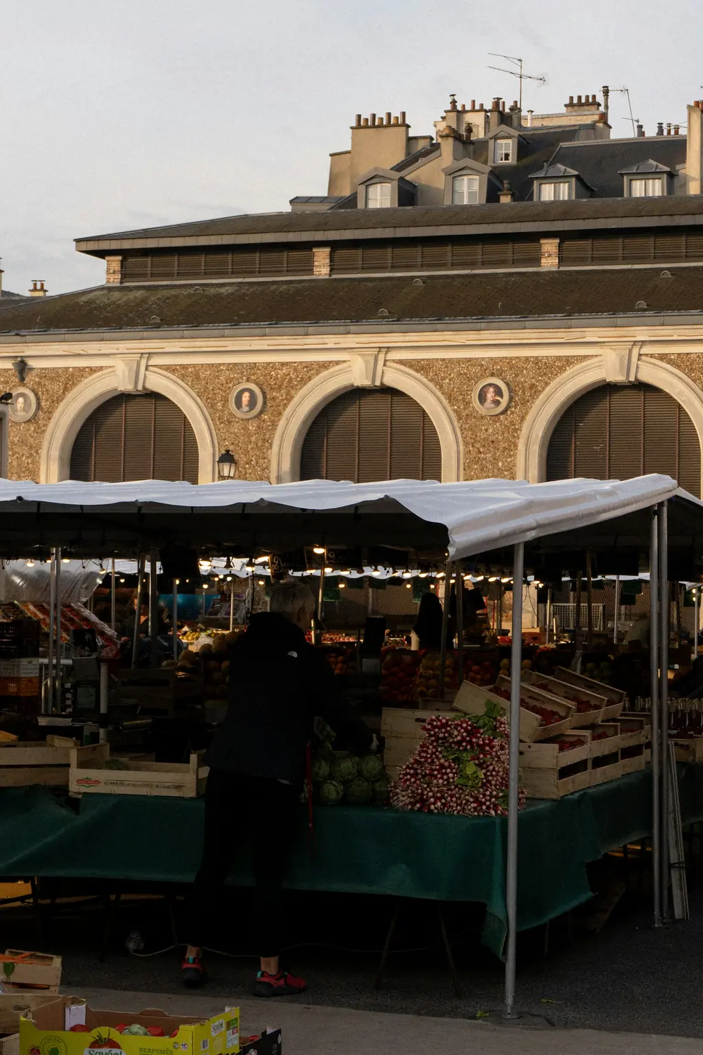 Étal de primeur sous un barnum éclairé, tôt le matin, devant une halle à arcades